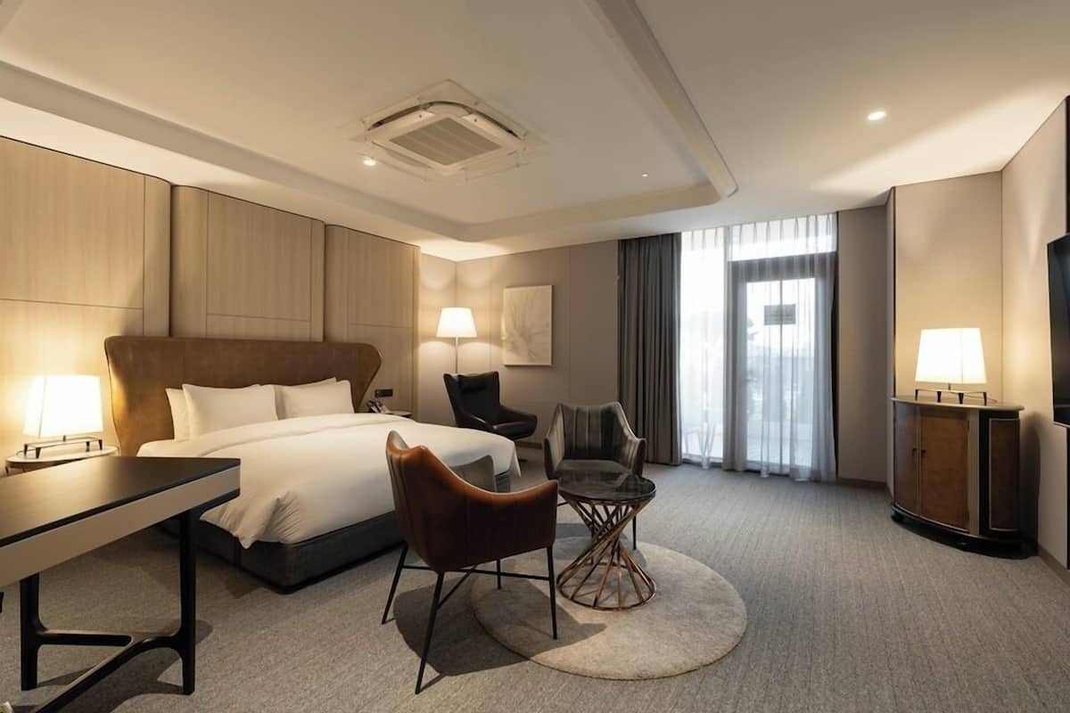 A modern hotel room with a large bed, a desk, two chairs, a round coffee table, a TV, and a large window with curtains.