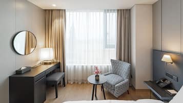 A hotel room with a bed, a desk with a lamp, a chair, a small table with a vase, and a phone on a desk.