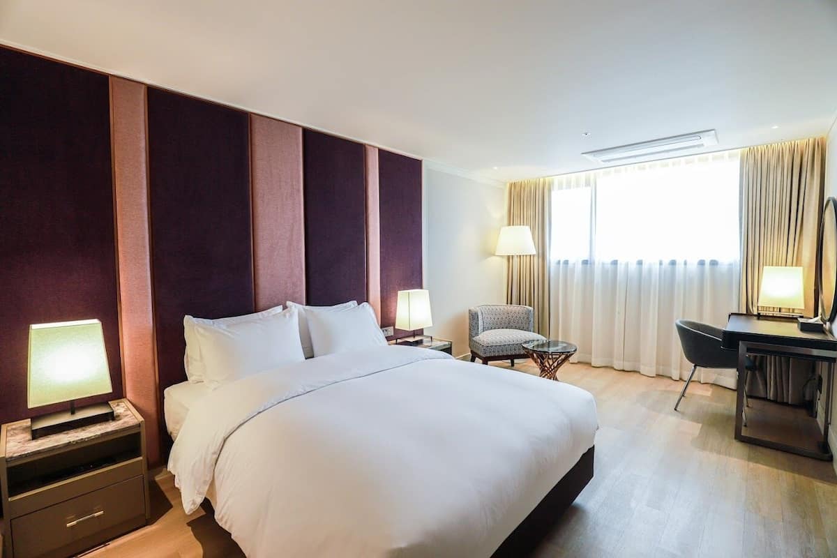 A hotel room with a large bed, two bedside tables with lamps, a desk with a chair, and a window with curtains.
