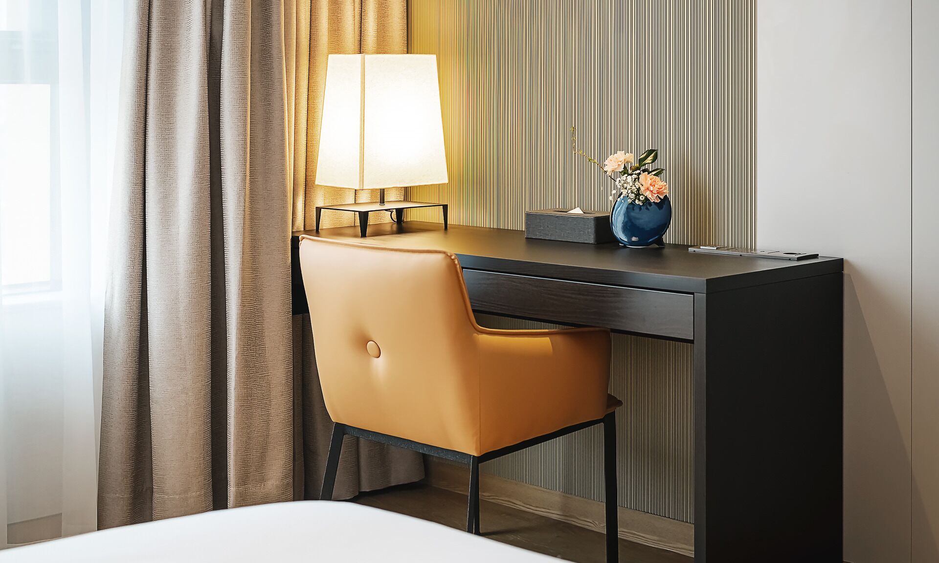 A hotel room with a desk, a chair, a lamp, and a vase with flowers.