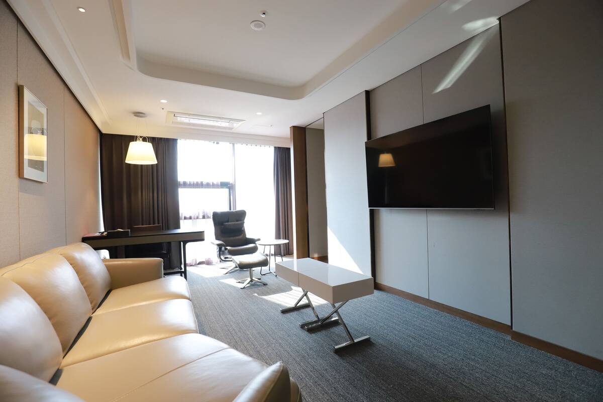 A modern hotel room with a leather sofa, a flat-screen TV mounted on the wall, a small table, and a chair.