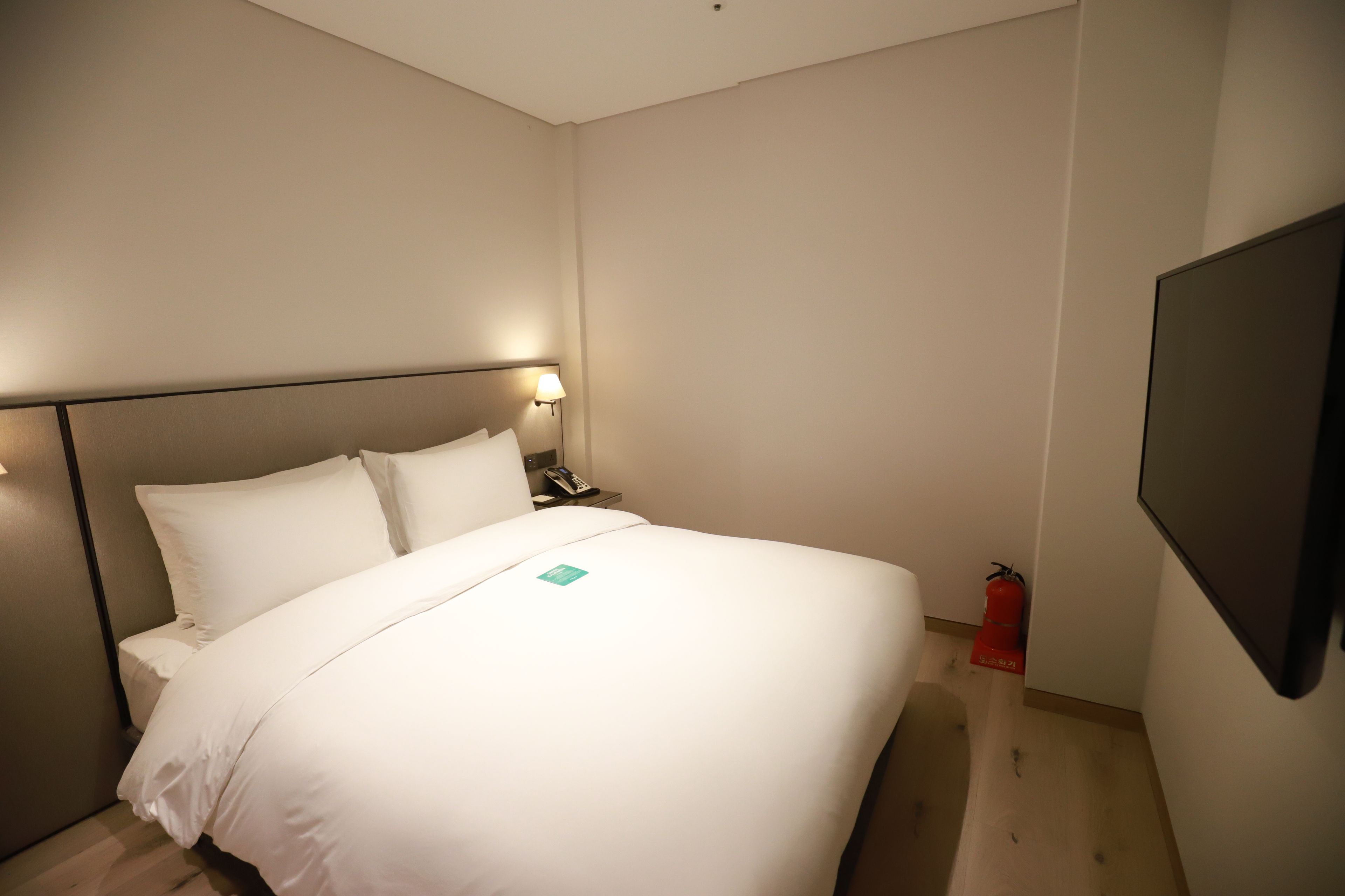 Habitación de hotel con cama, mesita de noche, lámpara y televisor de pared.