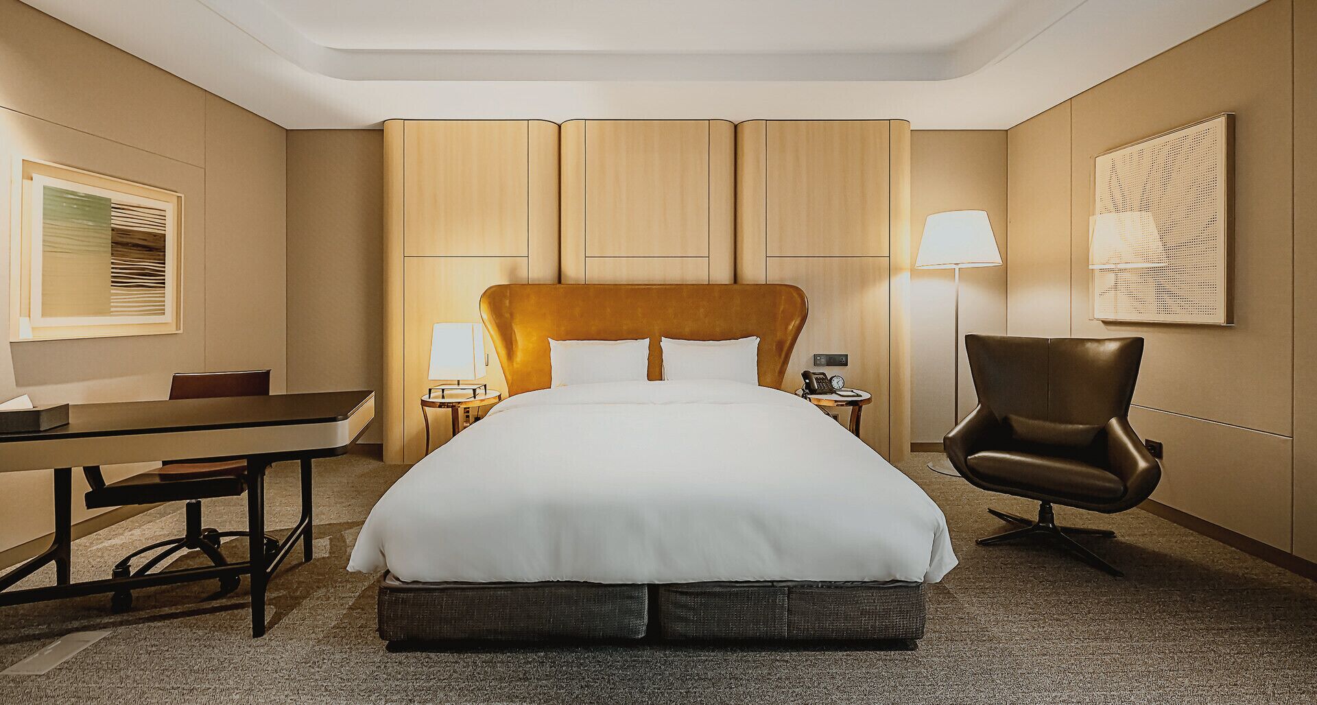 A hotel room with a large bed, a desk, a chair, a lamp, and a wall-mounted artwork.