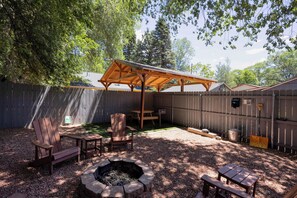 A backyard with a wooden pergola, two Adirondack chairs, a fire pit, and a picnic table.