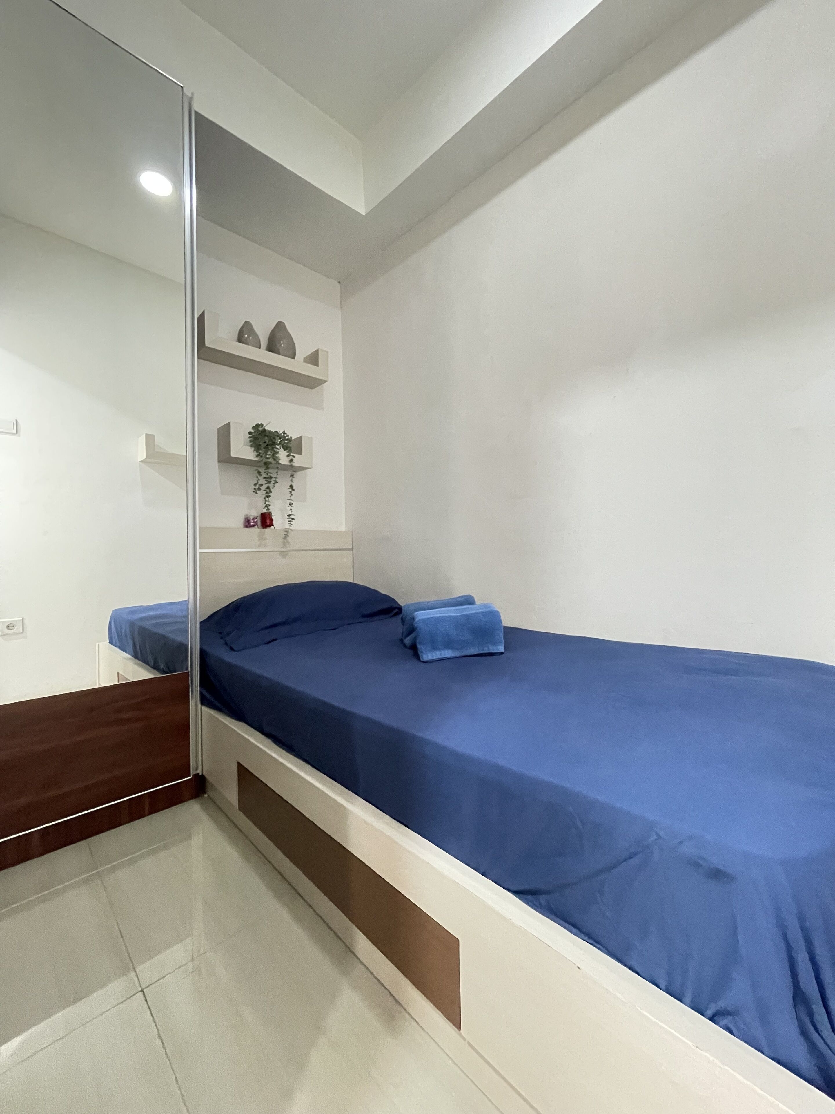 A compact bedroom with a single bed, a small shelf with decorative items, and a mirror.