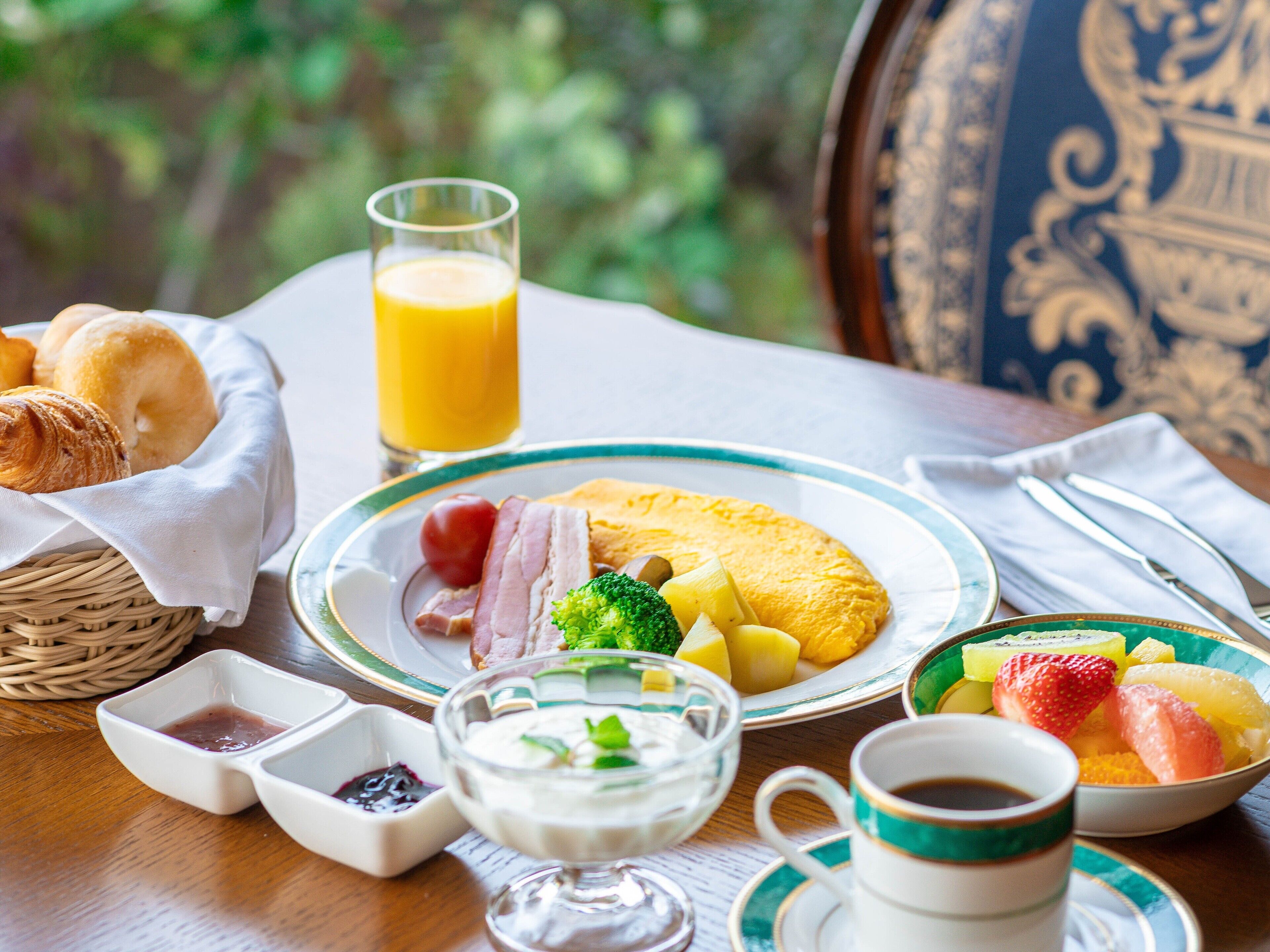 A breakfast setting with a plate of eggs, ham, and vegetables, a glass of orange juice, and a basket of croissants.
