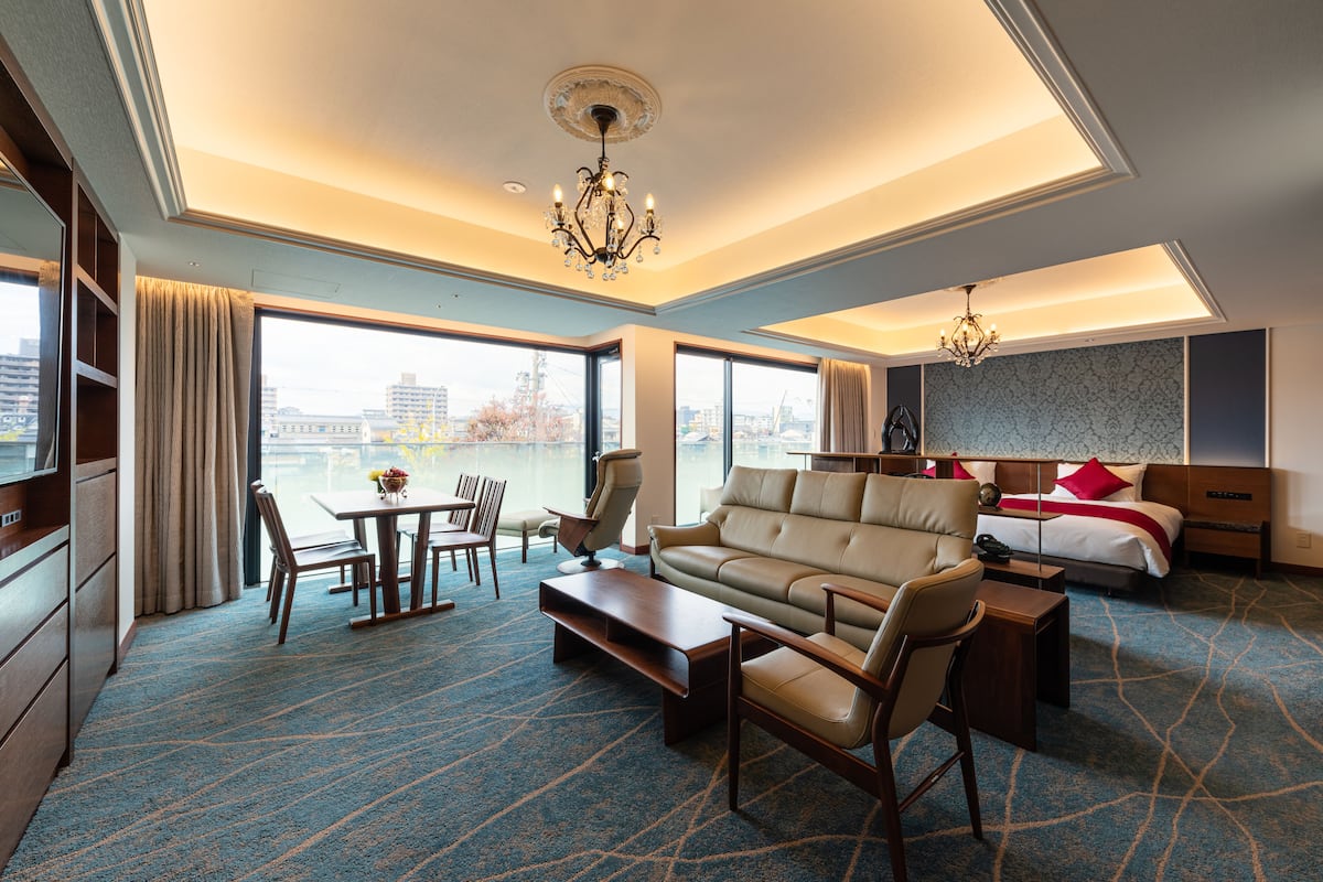 A hotel room with a large window, a sofa, a dining table, and a bed.