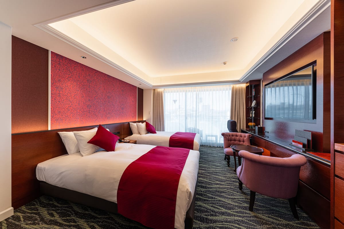 A hotel room with two beds, a desk, a chair, and a TV.