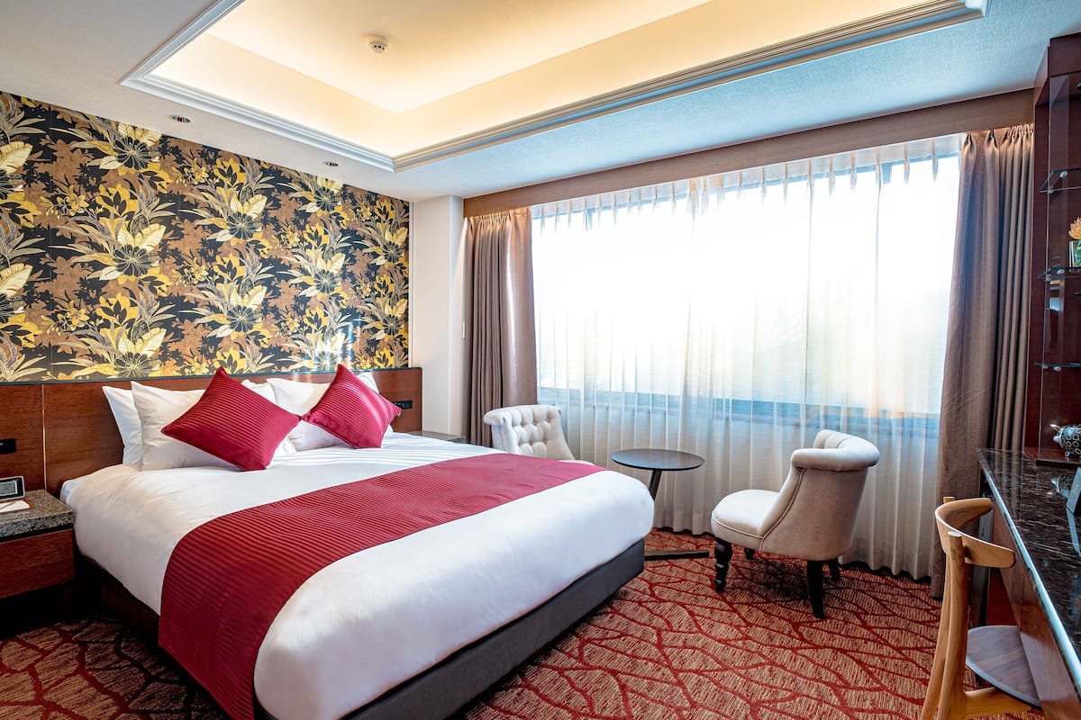 A hotel room with a large bed, two chairs, a small table, and a view of the sea through large windows.