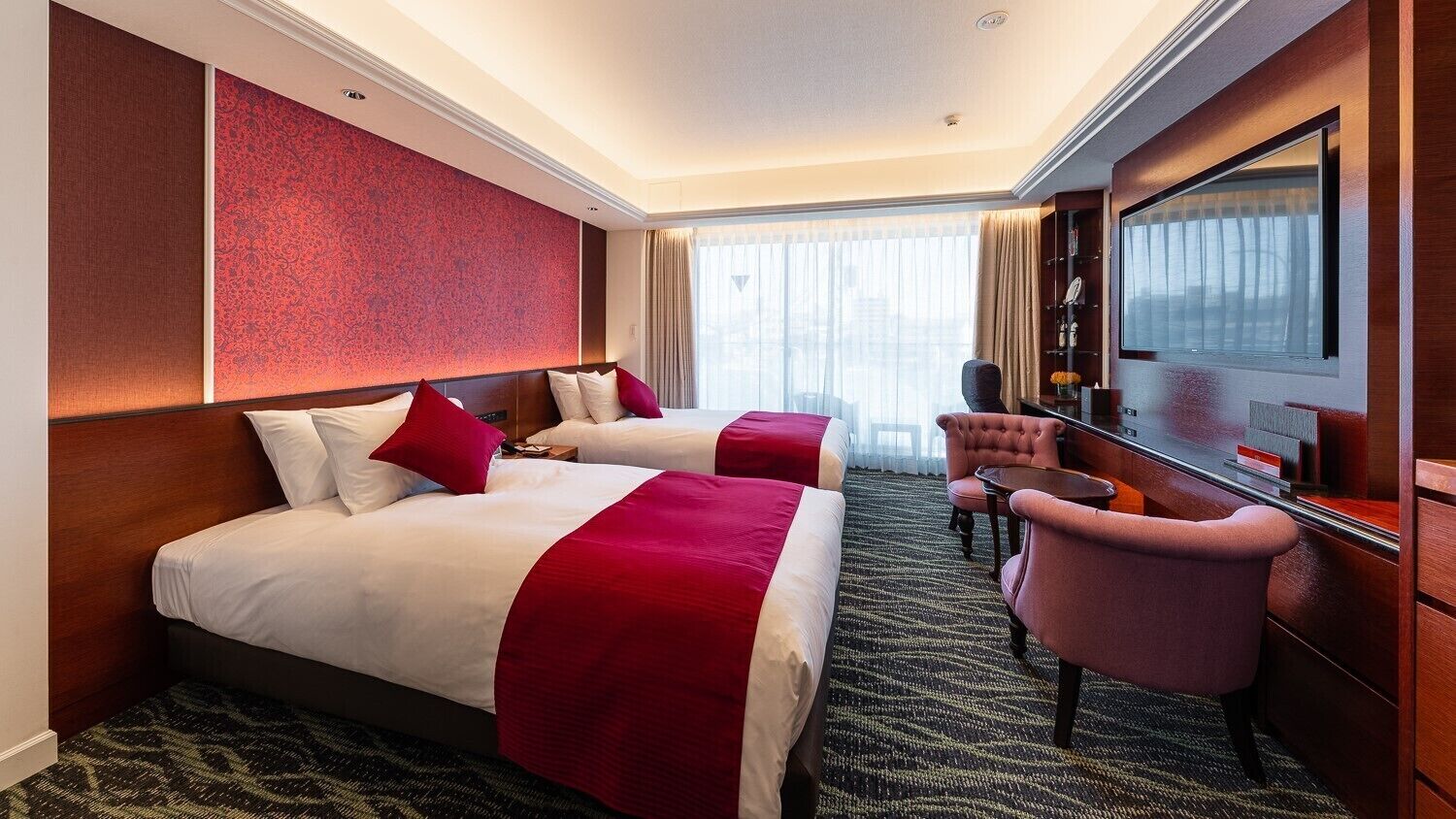 A hotel room with two beds, a desk, a chair, a television, and a large window.