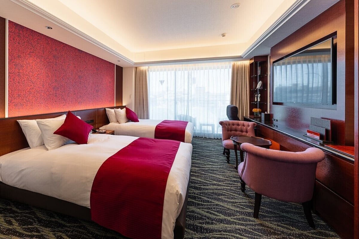 A hotel room with two beds, a desk, a chair, a television, and a large window.