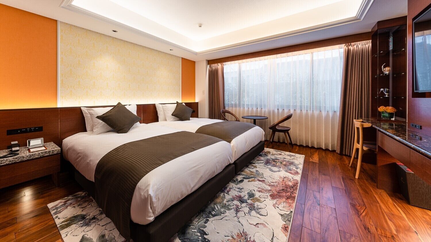 A hotel room with two beds, a desk, a chair, a TV, and a large window with curtains.