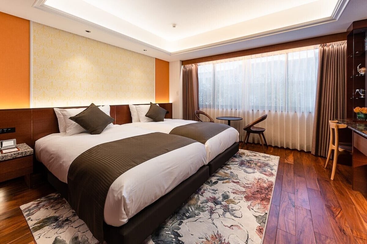 A hotel room with two beds, a desk, a chair, a TV, and a large window with curtains.