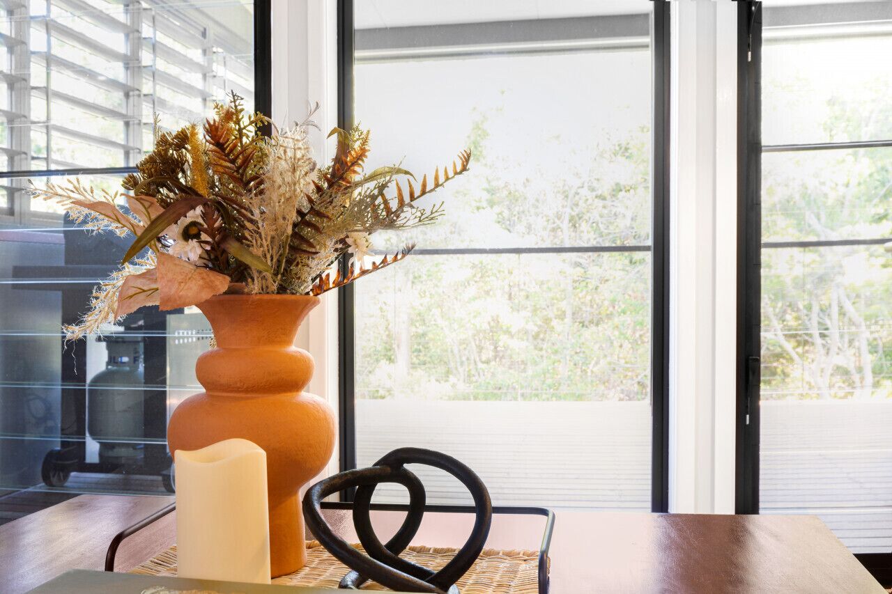 Eine Vase mit getrockneten Pflanzen auf einem Tisch neben einem Fenster mit Rollläden.