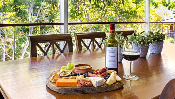 A wooden table with a charcuterie board, a bottle of wine, and a glass of wine on a balcony with potted plants and trees in the background.