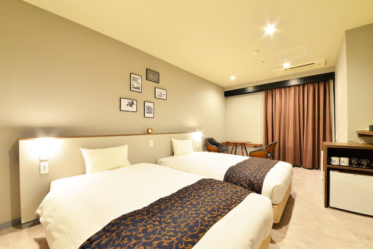A hotel room with two beds, a desk, and a chair. There are framed pictures on the wall and a window with curtains.