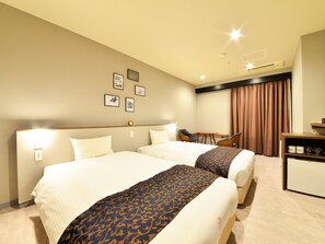 Ein Hotelzimmer mit zwei Betten, einem Schreibtisch und einem Sessel. An der Wand hängen gerahmte Bilder und es gibt ein Fenster mit Vorhängen.