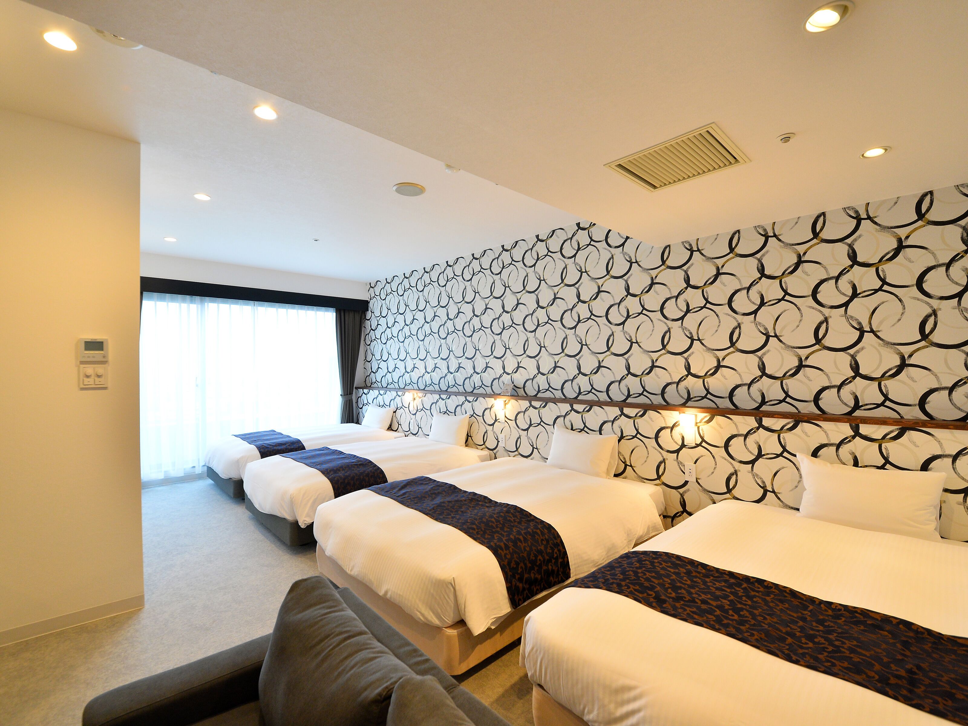 A hotel room with three beds, a large window with curtains, a patterned wall, and a ceiling with recessed lighting.