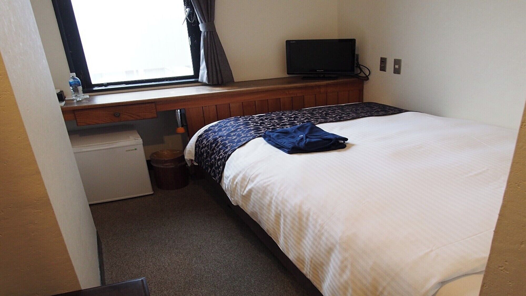 A bedroom with a bed, a television on a wooden shelf, a small refrigerator, and a window with curtains.