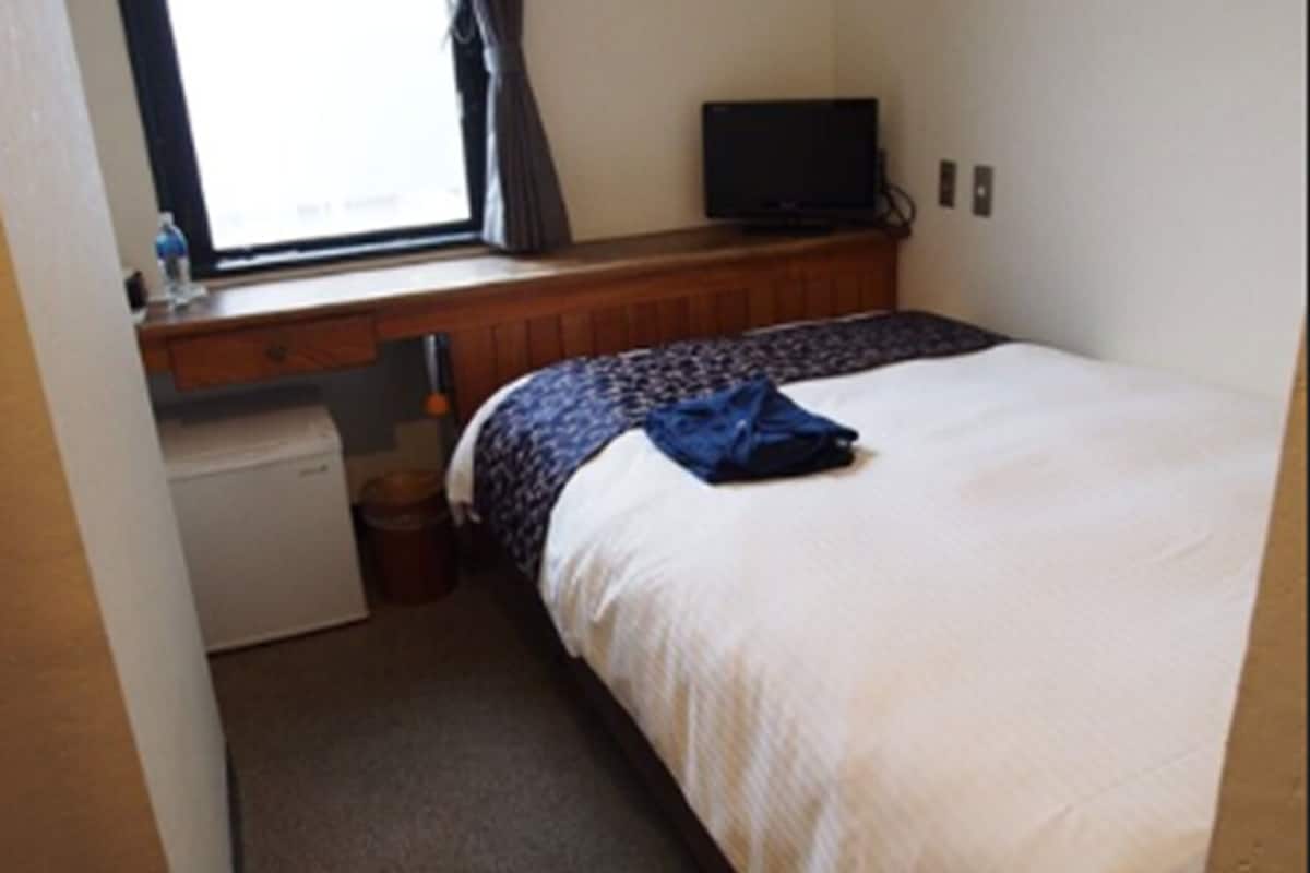 A bedroom with a bed, a small desk with a computer, a window with curtains, and a television on a stand.
