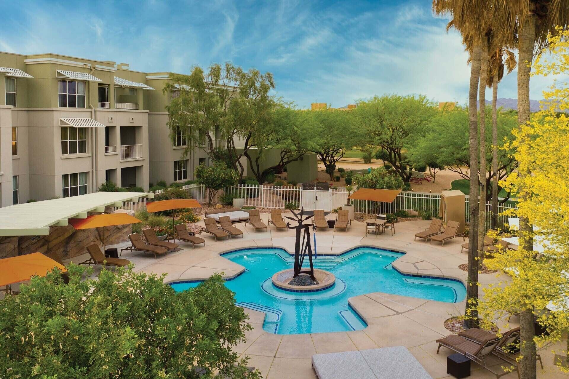A pool area with lounge chairs and a central water feature, surrounded by trees and a building.