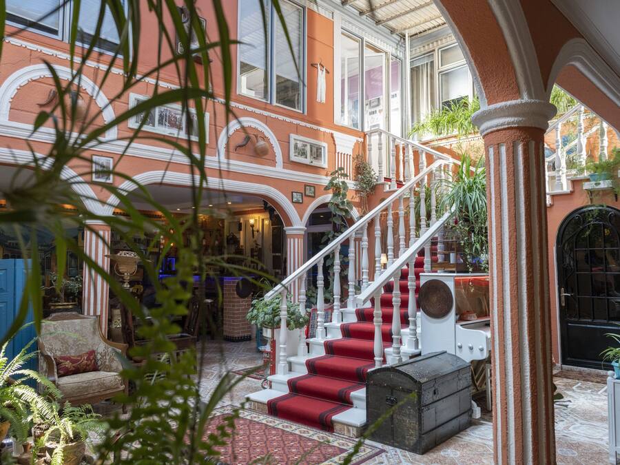A grand staircase with red carpeted steps leading to an upper floor, surrounded by ornate architecture and potted plants.
