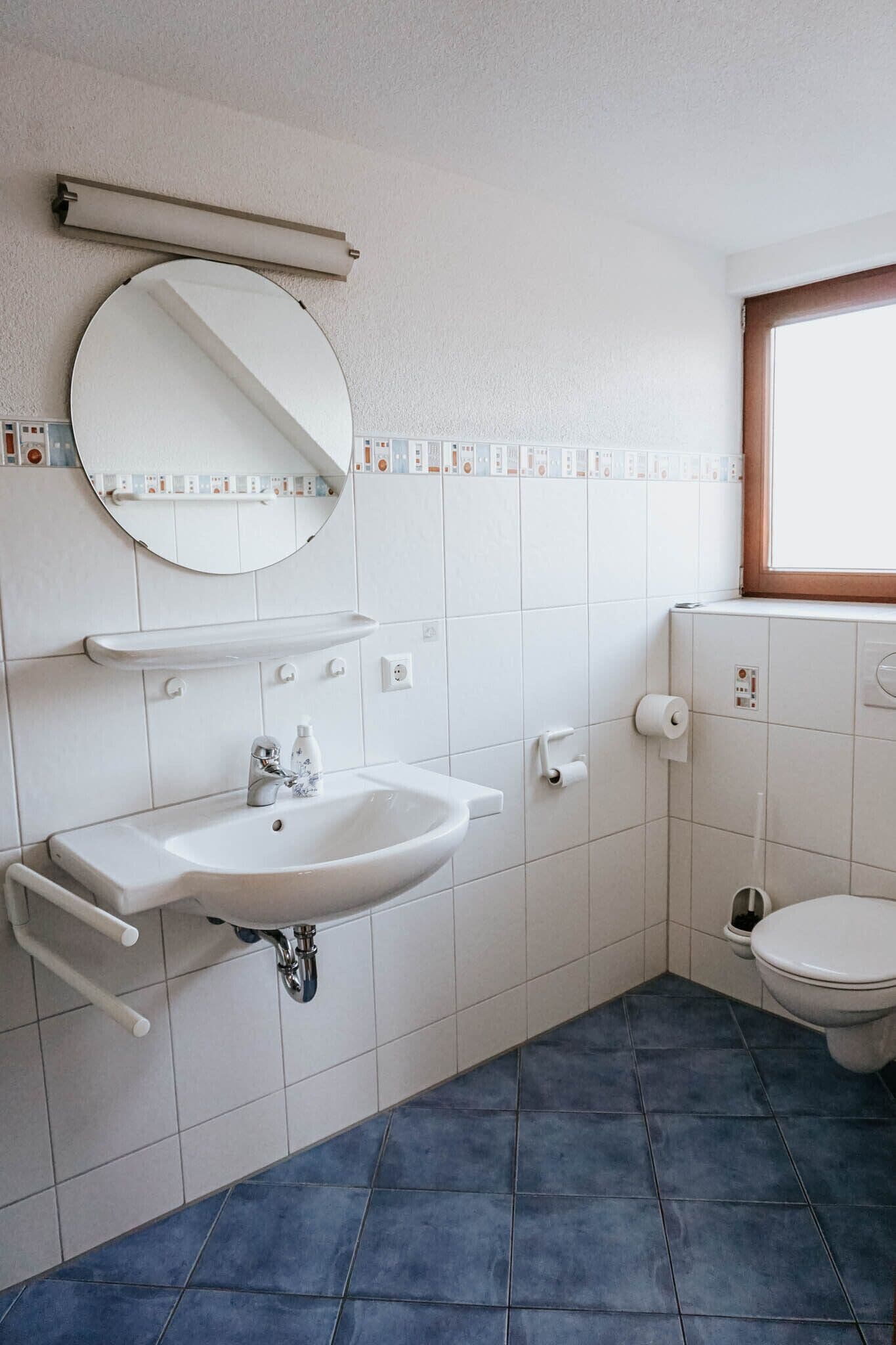 Un baño con lavamanos blanco, espejo y retrete, suelo de azulejos azules y una ventana con marco de madera.