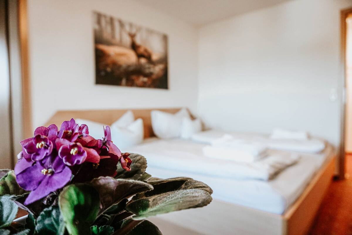 A neatly made bed with white linens and pillows, a framed picture on the wall, and a potted plant with purple flowers in the foreground.