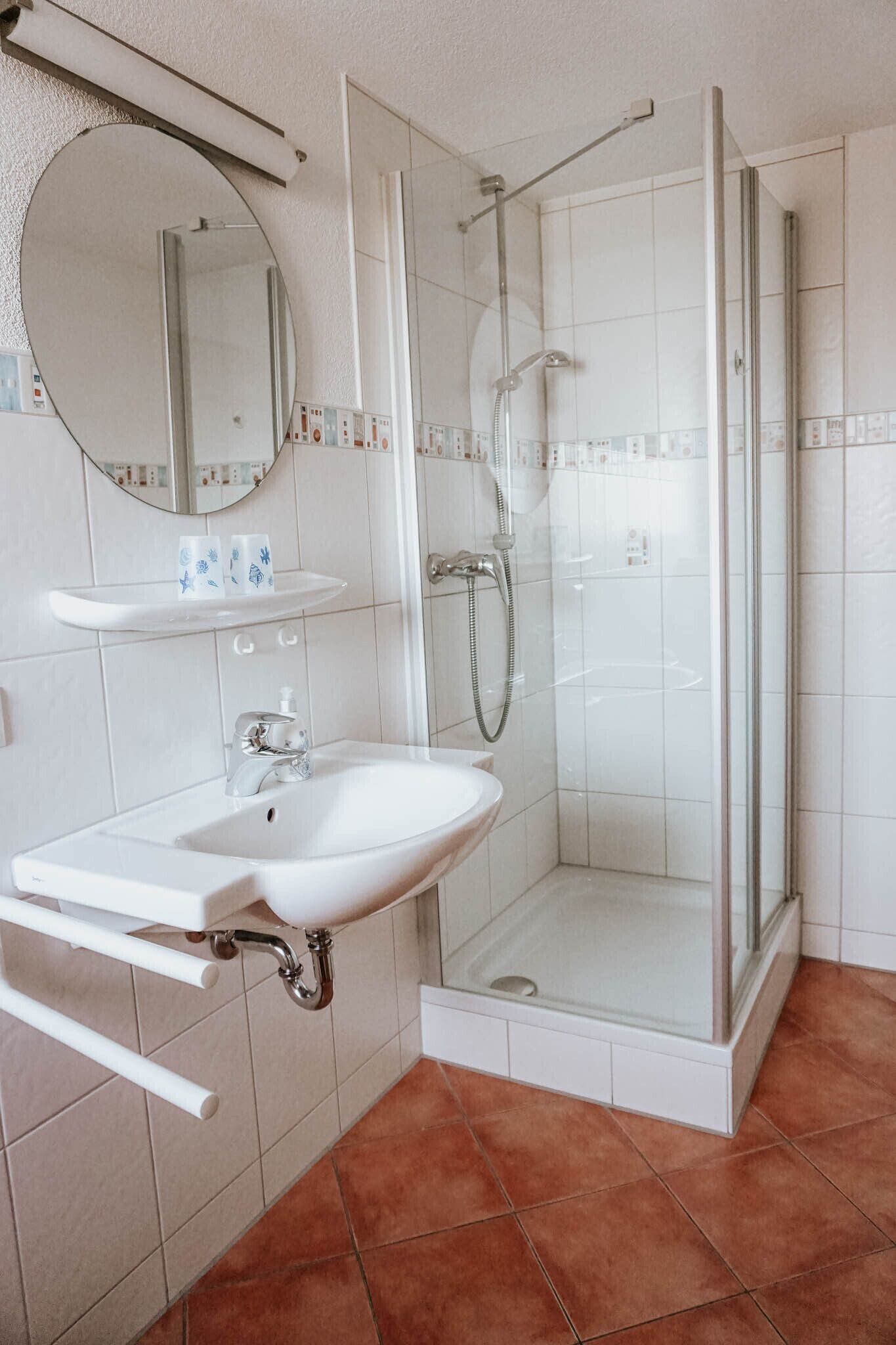 Ein Badezimmer mit einer gläsernen Duschkabine, einem weißen Waschbecken mit Spiegel darüber und einem gefliesten Boden.