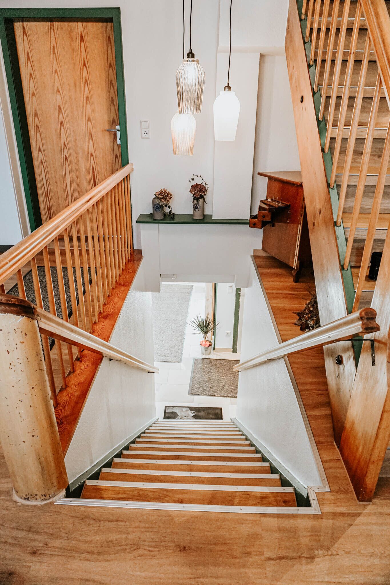 Un escalier en bois, avec une porte verte en haut, une petite table avec une plante et des lampes suspendues.