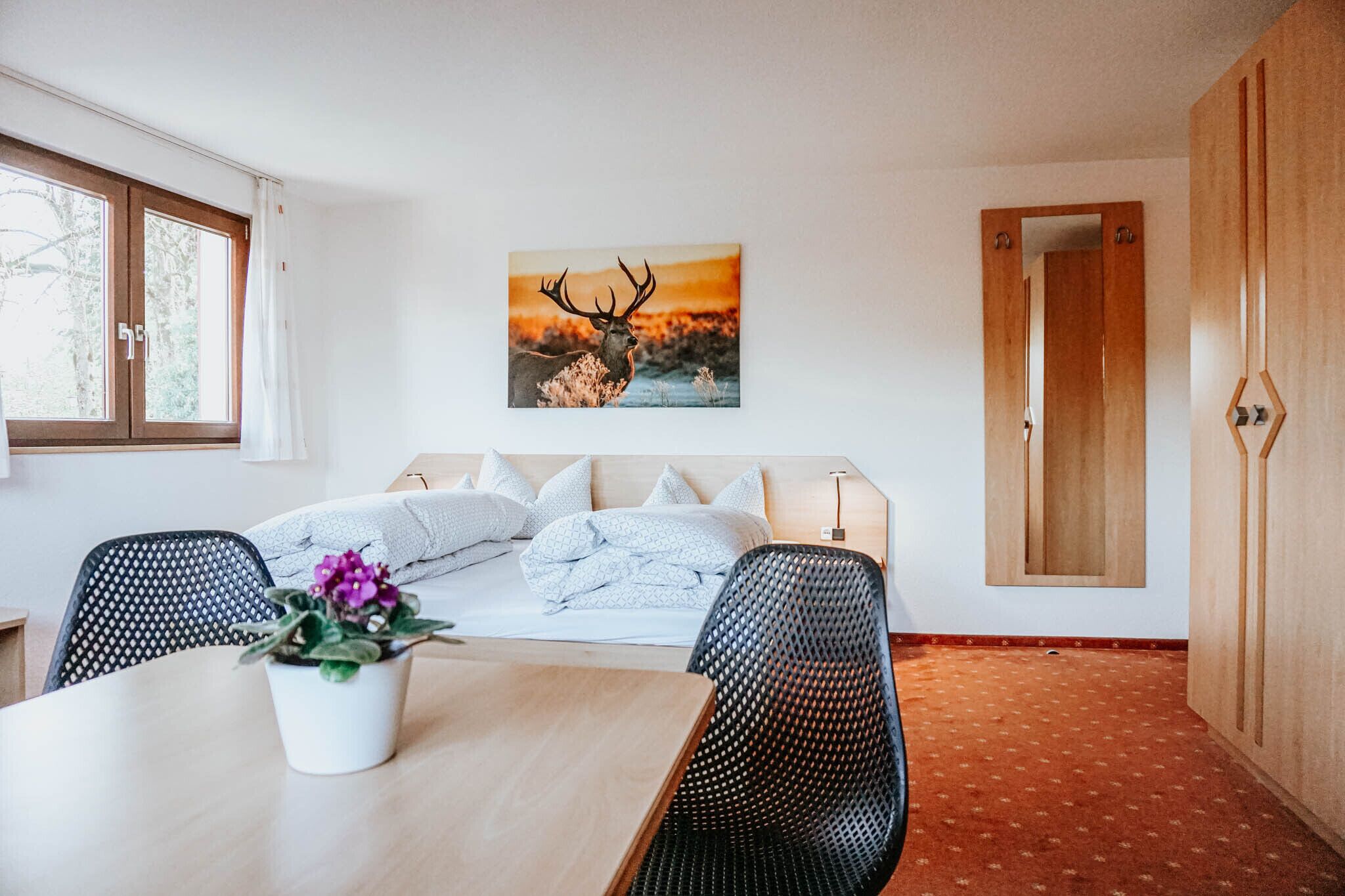 A bedroom with a bed, a dining table with chairs, a potted plant, and a painting of a deer.