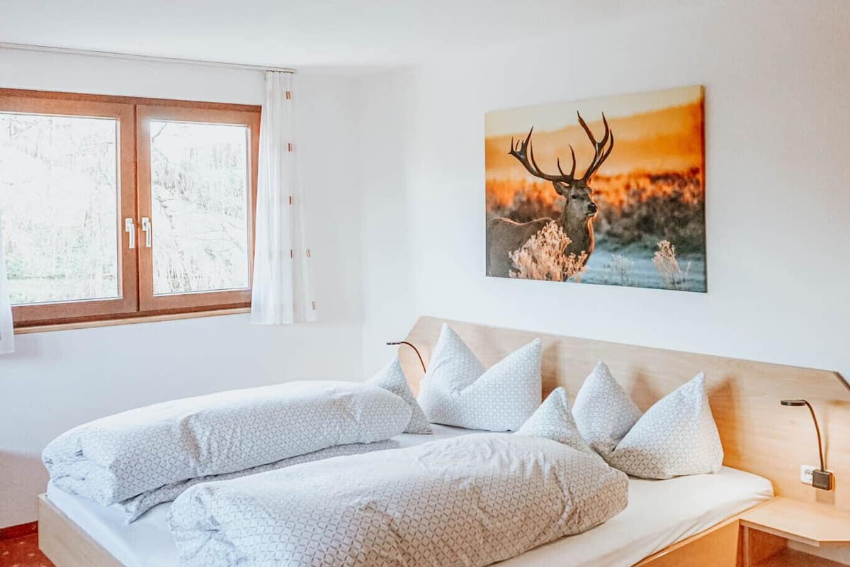 A bedroom with a wooden bed, white bedding, a window with curtains, and a deer painting on the wall.