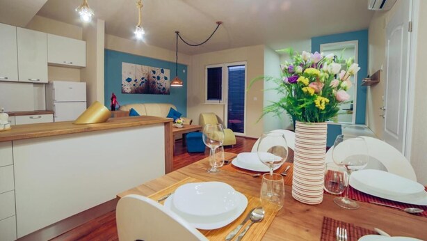 A dining area with a wooden table set for four, a vase of flowers, and a kitchen in the background.