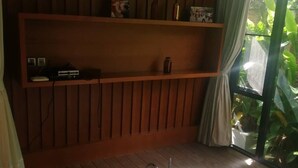 Une salle de bain avec un plancher en bois, une baignoire blanche, une étagère contenant divers objets et une fenêtre avec des rideaux.