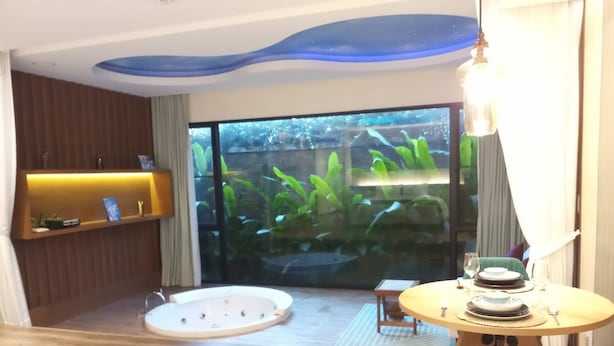 Une chambre d’hôtel moderne avec un mur en verre, une petite table ronde avec une chaise verte, un banc en bois et une étagère intégrée avec une lampe.