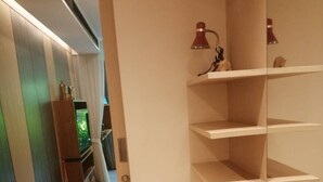 Une chambre moderne avec un bureau en bois, une chaise et un téléviseur posé sur un meuble. Des étagères sont fixées au mur, et une lampe est visible.