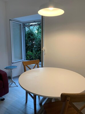 A room with a round table and two wooden chairs, a small side table, and a window with a view of greenery outside.