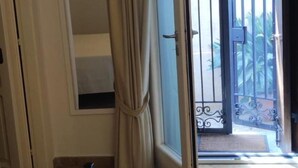 A hotel room with a bed, a desk, a chair, a window with curtains, and an open door leading to an outdoor area with a gate. - Locanda Badia Residenze Iride e Celeste (Castellabate)