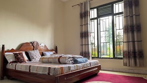 A bedroom with a wooden bed, patterned bedding, a red carpet, and a window with curtains. - Room in B&B - Nice Rooms, Unbeatable Prices (Muhoza)