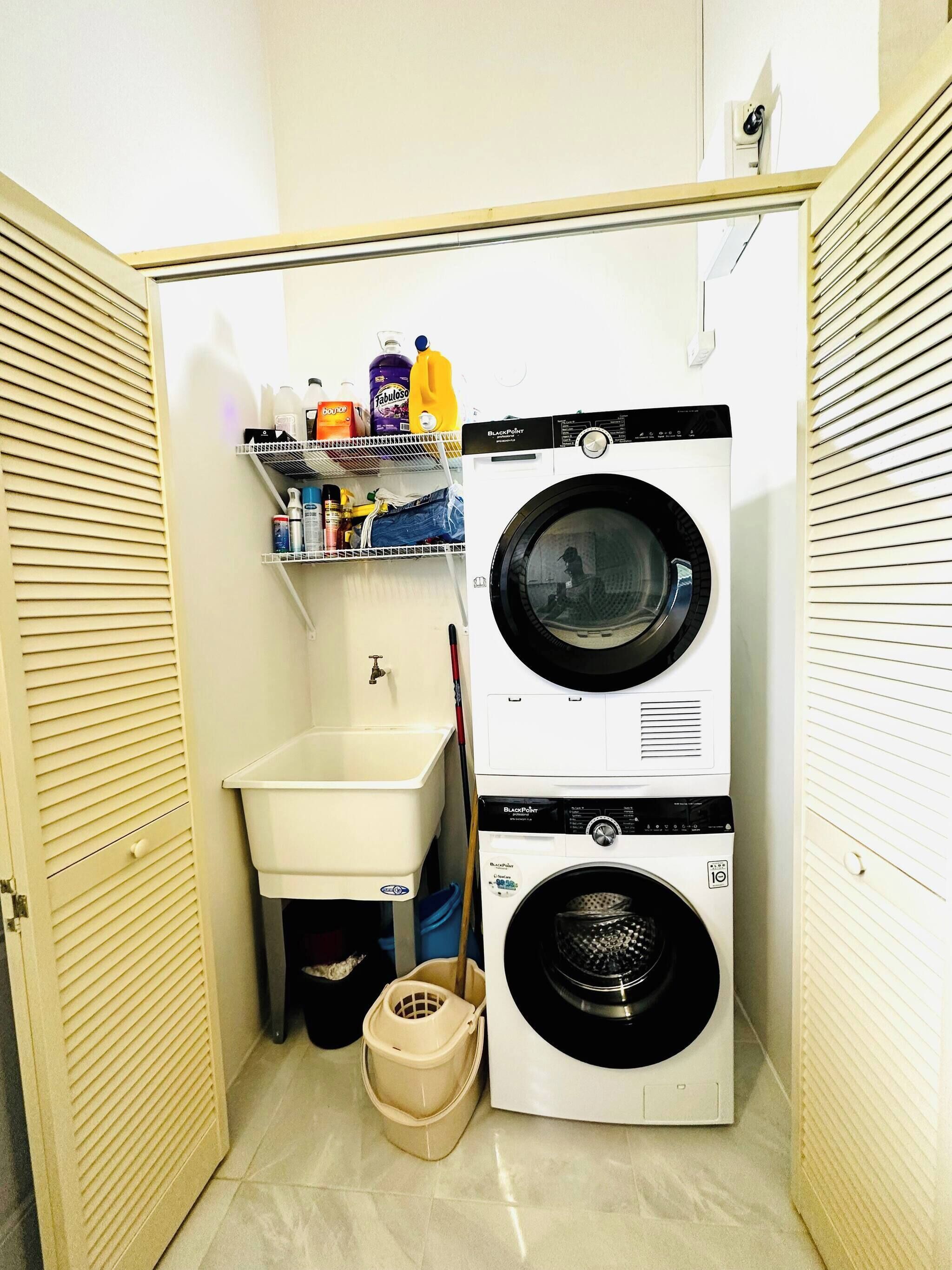 Une petite buanderie équipée d’un lave-linge et d’un sèche-linge superposés, d’un petit évier et d’étagères contenant des produits de nettoyage.