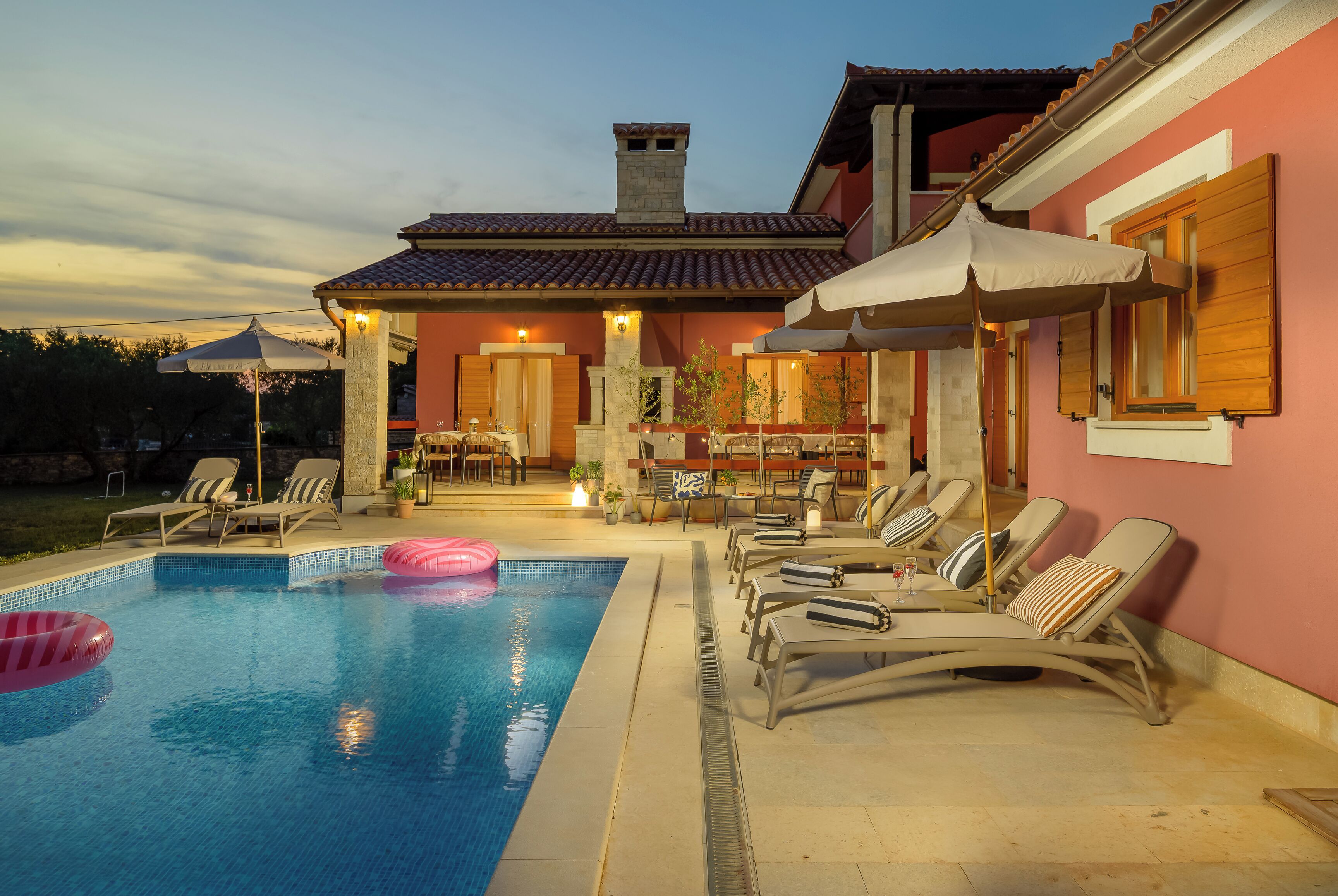 A swimming pool with lounge chairs and umbrellas, a patio area with outdoor seating, and a building with a tiled roof.