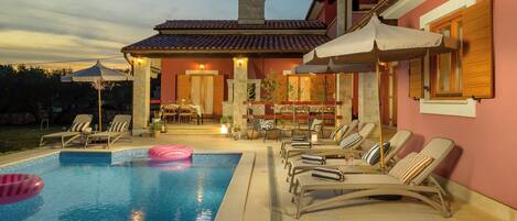 A swimming pool with lounge chairs and umbrellas, a patio area with outdoor seating, and a building with a tiled roof.