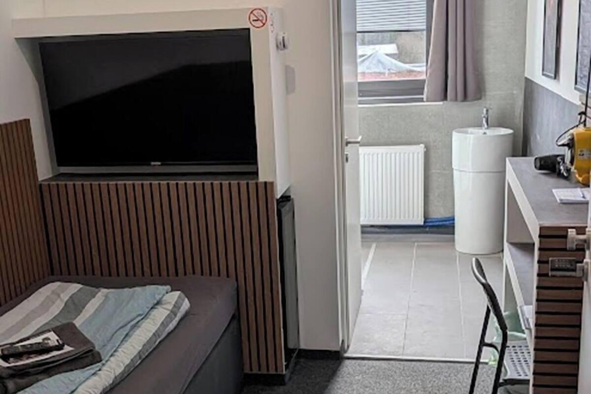 A compact hotel room with a bed, a TV, a desk, and a bathroom visible through an open door.