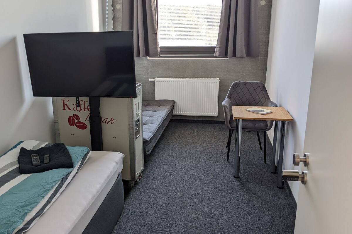A compact hotel room with a single bed, a small desk, a chair, a television, and a window with curtains.