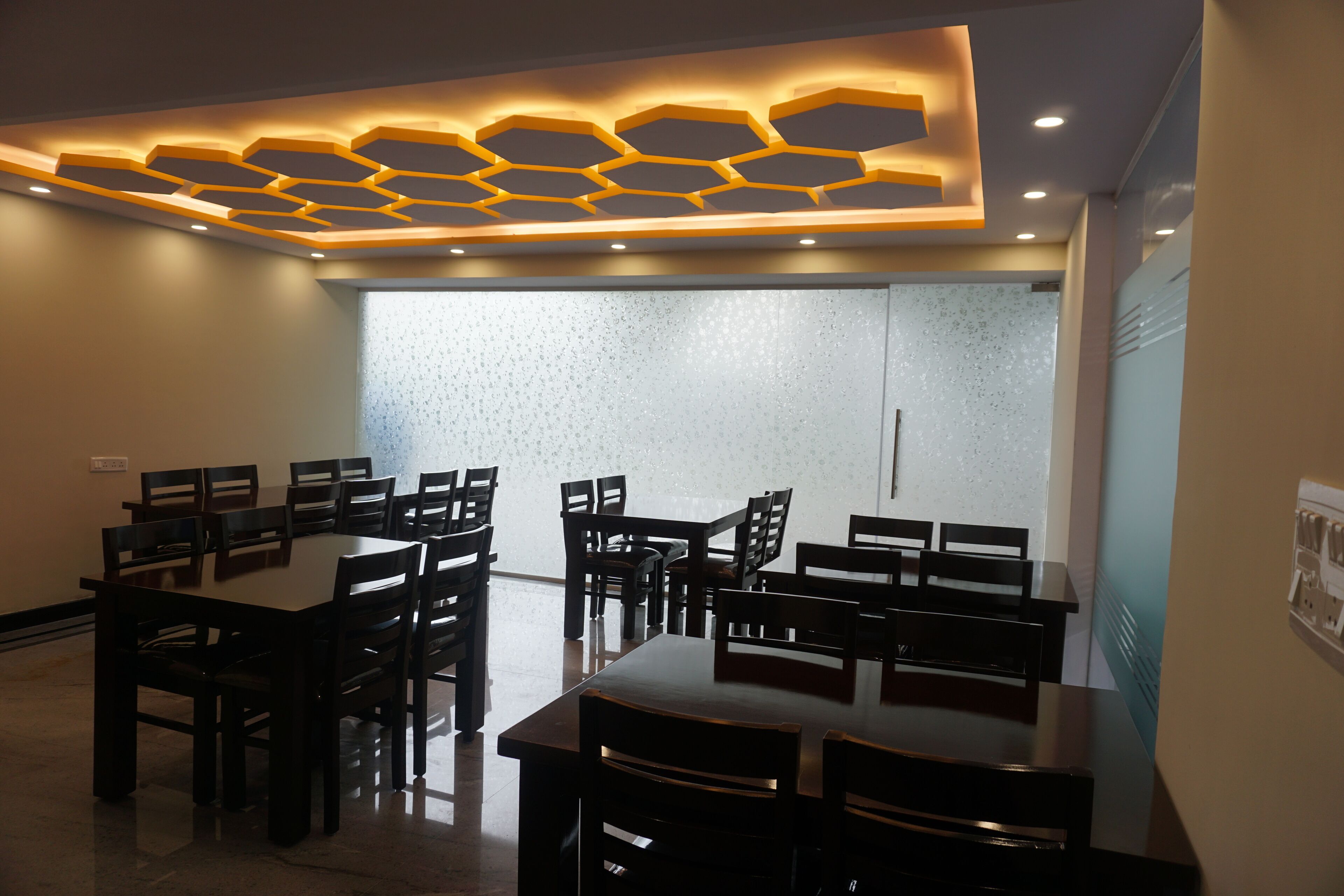 A conference room with a large table and multiple chairs, a ceiling with hexagonal light fixtures, and a frosted glass partition.