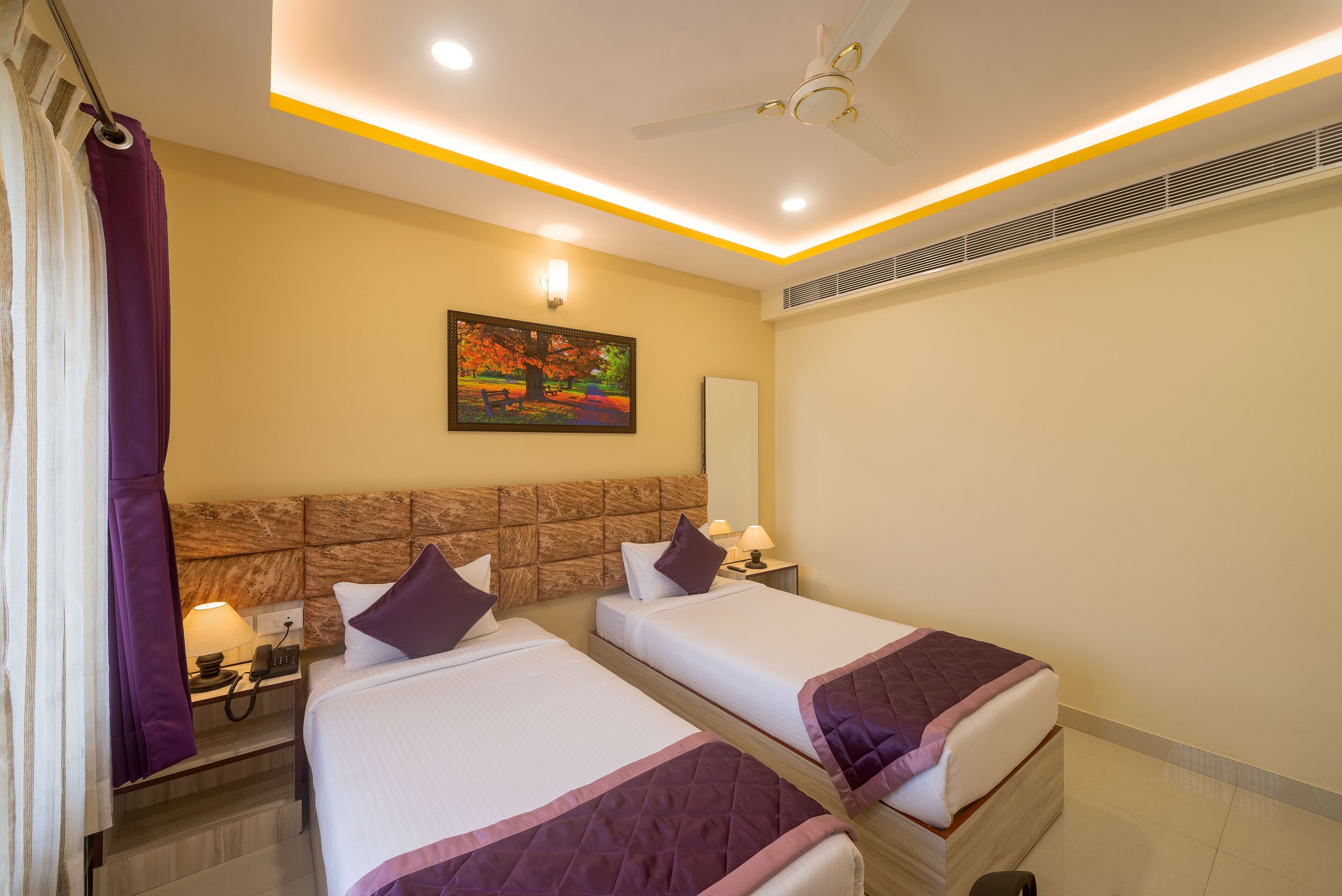 A hotel room with two beds, a painting on the wall, a ceiling fan, and a nightstand with a lamp.