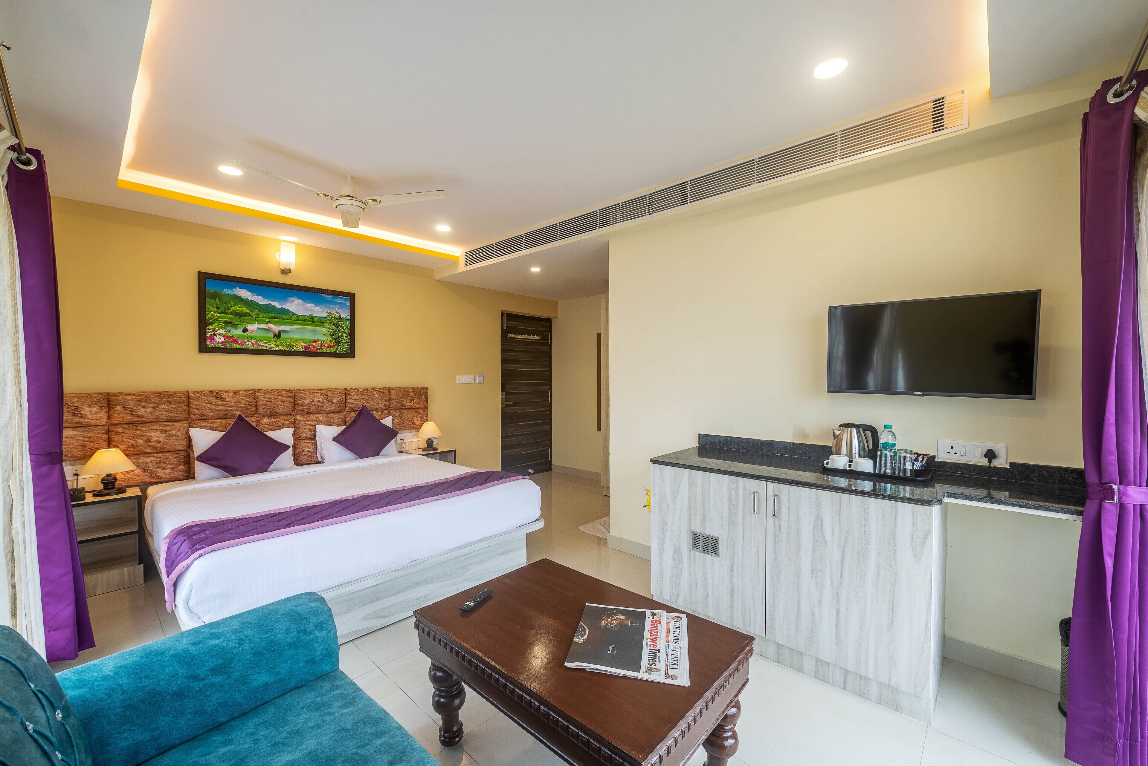 A hotel room with a bed, a sofa, a coffee table, a TV, and a kitchen area.
