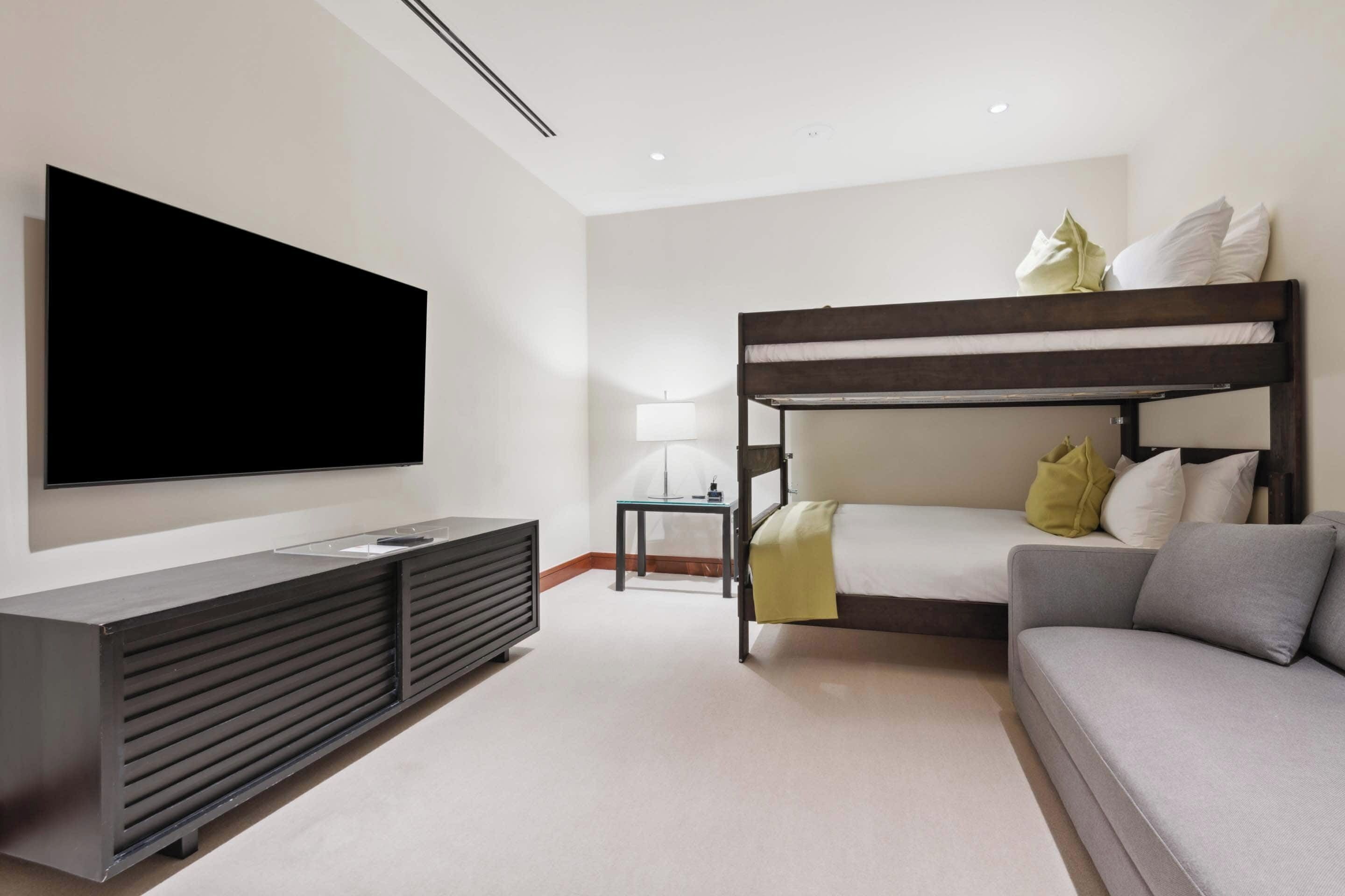 A modern hotel room with a bunk bed, a flat-screen TV, a sofa, and a minimalist design.