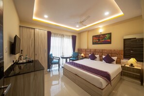 A hotel room with a bed, a desk with a chair, a TV, a painting, and a ceiling fan. - Zenith Hotels Hebbal Bangalore (Bengaluru)