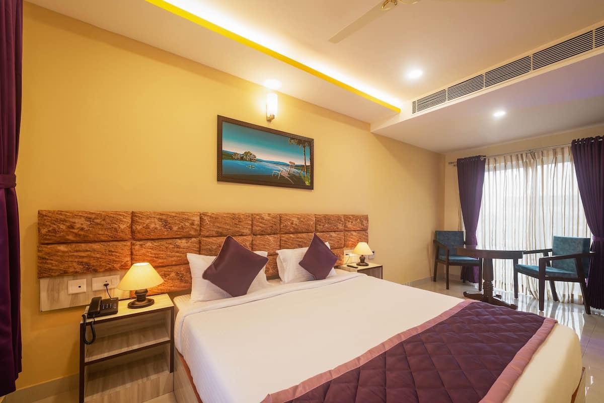 Quarto de hotel com uma cama grande, mesa de cabeceira com abajur, quadro na parede, pequena mesa com cadeiras e janela com cortinas.